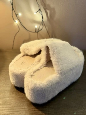 Stand Studio platåtofflor - Trendy beige tofflor från Stand Studio med fluffig pälsimitation och stor platåsula. Tofflorna har öppen tå och en bred rem över foten. Sparsamt använda. Coola både på fötterna och som inredningsdetalj 💅🏼