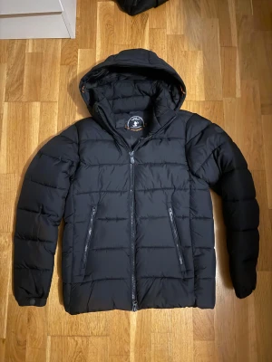 Save the Duck jacka  - Passar som S. Säljer en svart pufferjacka från Save The Duck med huva och dragkedja. Jackan är tillverkad av återvunnen polyester och har Plumtech-fyllning som är helt animal free. Snygg, stilren och perfekt för kalla dagar.