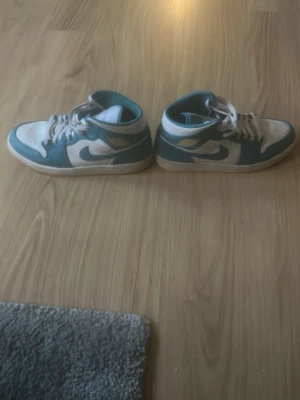 Nike Jordan 1 Mid blå och vita sneakers - Säljer ett par Nike Jordan 1 Mid sneakers i blått och vitt med klassisk swoosh på sidan. Skorna har snörning, rund tå och vit sula med gummiyttersula. Perfekta för dig som gillar streetwear och vill ha en ikonisk look.