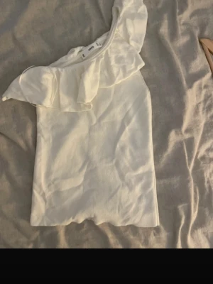 Vit oneshoulder topp med volang - Snygg vit oneshoulder topp med bred volangdetalj över axeln. Perfekt för dig som vill ha en unik och trendig look. Tunn och lätt i materialet, passar till många olika stilar.