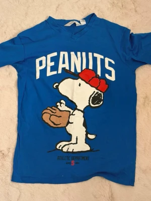 Blå Peanuts t-shirt med Snoopy💙 - Blå t-shirt med stort tryck av Snoopy i röd keps och basebollhandske på framsidan samt texten 'PEANUTS'. På baksidan står det '50' med Woodstock och en baseboll. T-shirten har v-ringning och korta ärmar. Perfekt för dig som gillar klassiska serier och sportig stil!💙