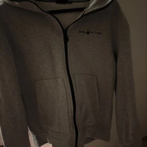 Grå hoodie från Sail Racing - Grå tjock zip hoodie från Sail Racing. Använd fåtal gånger, har dock en liten fläck på ryggen.