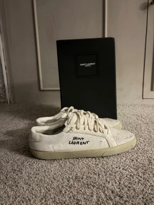 Vita Saint Laurent sneakers - Säljer ett par vita Saint Laurent sneakers i canvas med handskriven logga på sidan. Skorna har snörning, rund tå och platt sula. Klassisk och stilren design som passar till det mesta. Kommer med originalkartong. Priset kan diskuteras 