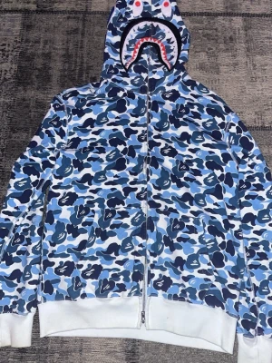 BAPE blå camo shark hoodie - Säljer en ikonisk BAPE shark hoodie med blått camomönster och dragkedja hela vägen upp. Huvan har hajmotiv med tänder och ögon, samt broderade bokstäver på sidan. Ribbstickade muddar och fickor framtill. Tillverkad i mjuk bomull för maximal komfort.