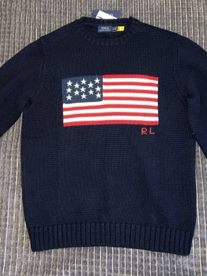 Mörkblå stickad tröja Polo Ralph Lauren - Mörkblå stickad tröja från Polo Ralph Lauren med amerikansk flagga i rött, vitt och blått på bröstet samt RL-broderi. Tröjan har rund halsringning och ribbade muddar vid ärmslut och nederkant. Perfekt för dig som gillar klassisk preppy stil.