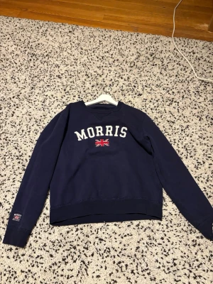 Mörkblå sweatshirt från Morris - Mörkblå sweatshirt från Morris med vit broderad logga och brittisk flagga på bröstet. Tröjan har rund halsringning, ribbade muddar och en liten patch på ärmen. Perfekt för en avslappnad och stilren look.
