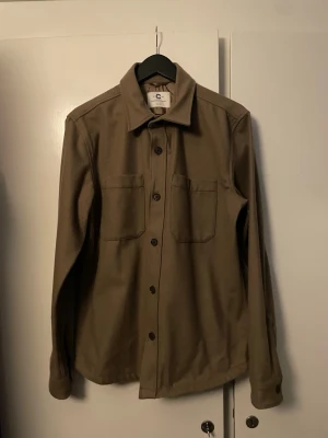 Beige overshirt med knappar - Stilren beige overshirt med klassisk krage och två stora bröstfickor. Långärmad modell med knäppning framtill och raka ärmslut. Perfekt att bära som lager över t-shirt eller hoodie för en avslappnad look. Endast använts en gång 