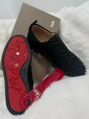 Svarta Louboutin sneakers med nitar - Säljer ett par svarta sneakers från Christian Louboutin med ikonisk röd sula och svarta nitar på tån. Skorna har snörning och är tillverkade i skinn och mocka för en lyxig känsla. Perfekta för dig som vill sticka ut med en edgy och exklusiv stil.