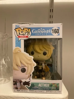 Funko Pop Genshin Impact Aether - Säljer en Funko Pop vinylfigur av Aether från Genshin Impact, nummer 160. Figuren har blont hår och detaljerade kläder i svart, vitt och guld. Levereras i originalförpackning, perfekt för samlare eller fans av spelet.