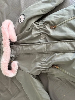 Grön parkas med rosa fuskpäls - Stilren grön parkas med mjuk rosa fuskpäls runt huvan och coolt 'Girls League'-märke på ärmen. Jackan har dragkedjor framtill och markerad midja för snygg passform. Perfekt för kalla dagar när du vill vara både varm och trendig.
