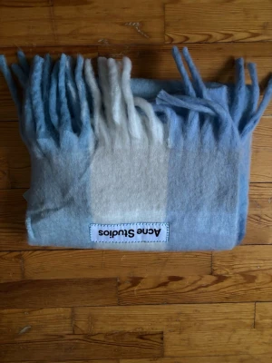 Ljusblå och vit halsduk Acne Studios - Mjuk och fluffig halsduk från Acne Studios i ljusblått och vitt med breda blockränder och långa fransar. Tillverkad i ullblandning och har en tydlig Acne Studios-logga påsydd. Perfekt accessoar för att lyfta din outfit.