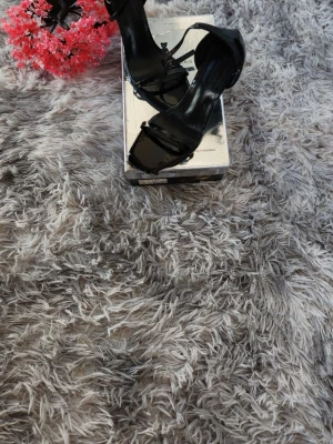 Svarta lackade pumps med hög klack - Säljer ett par svarta pumps i lackat material med öppen tå och hög klack. Skorna har en elegant och stilren design med smala remmar över foten och runt ankeln. Perfekta för dig som vill ha en klassisk och trendig look.