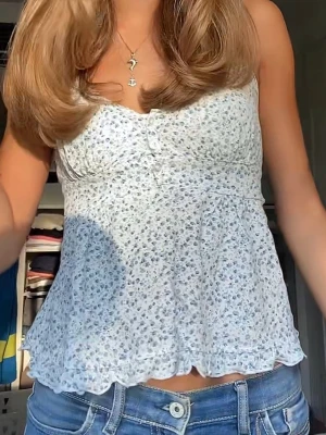Blommig vit blus/topp med knappar - Söt blommig blus i vitt med små blå och vita blommor. Toppen har en lätt utsvängd passform, knappar framtill och volangkant nedtill. Perfekt för sommaren. Köpt på brandy Melville för längesedan så den finns inte kvar och är väldigt fin. 