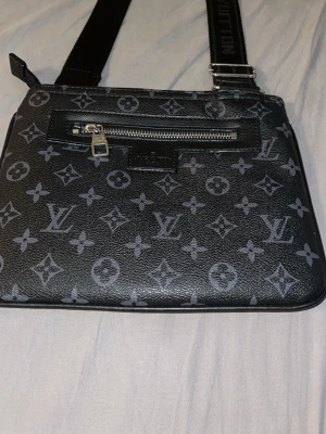  svart axelväska bytes kan!!!!! - Säljer en svart axelväska  med klassiskt monogrammönster i grått. Väskan har justerbar axelrem, en framficka med dragkedja och detaljer i silverfärg. Tillverkad i skinnliknande material och perfekt för dig som vill ha en stilren och ikonisk accessoar.