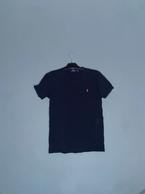Mörkblå t-shirt från Polo Ralph Lauren ej äkta - Snygg mörkblå t-shirt från Polo Ralph Lauren (ej äkta) med klassisk röd broderad logga på bröstet. T-shirten har rund halsringning och korta ärmar. Tillverkad i mjuk bomull som känns skön mot huden. Perfekt till jeans eller shorts för en clean look.