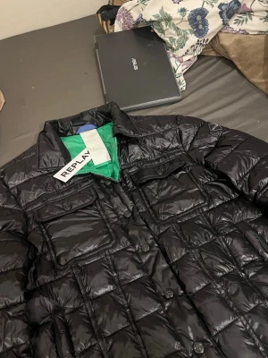 Svart dunjacka från Replay - Snygg svart dunjacka från Replay med quiltad design och knappar framtill. Jackan har två bröstfickor och en grön insida som ger en cool kontrast. Perfekt för dig som vill ha en stilren och modern look under kalla dagar.