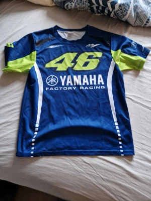 Yamaha fransk stil tshirt - Snygg blå t-shirt från Yamaha Factory Racing med stort nummer 46 i neongult på bröstet och vita detaljer. T-shirten har gröna partier på ärmarna och tryckta loggor. Perfekt för er son gillae franska stilen. Endast använd en gång.