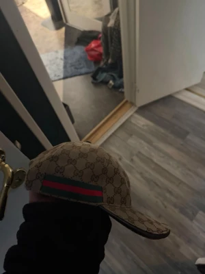 Gucci keps - Säljer en beige keps från Gucci med klassiskt GG-monogram över hela kepsen och en grön-röd rand på sidan. Kepsen har böjd skärm och justerbar rem bak. Insidan är fodrad i svart tyg och har Gucci-taggar. Perfekt för dig som gillar lyxiga accessoarer.
