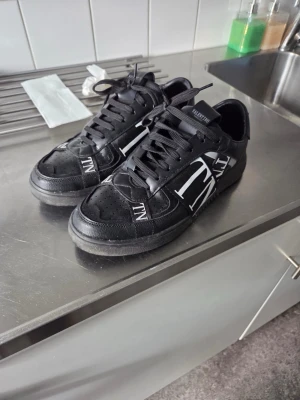 Svarta Valentino sneakers med vit logga - Säljer ett par svarta sneakers från Valentino med stor vit VLTN-logga på sidan. Skorna har snörning, rund tå och är tillverkade i skinn med en platt sula. Perfekta för dig som gillar stilrena och exklusiva sneakers. Äkta med serienummer i plösen. Inklusive lådan och dustbag samt påsen från vallentino store.