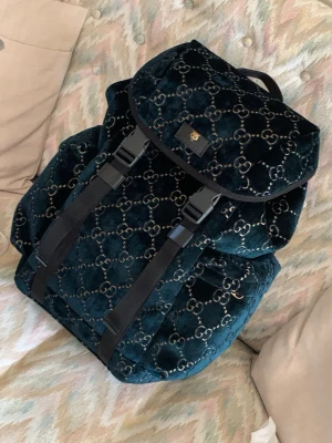Mörkblå sammet ryggsäck från Gucci - Lyxig mörkblå ryggsäck från Gucci i mjuk sammet med klassiskt GG-mönster i silver. Ryggsäcken har justerbara axelband i läder, praktiskt lock med spännen och rymligt huvudfack. Perfekt för dig som vill ha en statement-väska med exklusiv känsla.