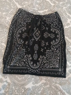 Svart strandkjol med bandanamönster - Snygg svart strandkjol med vitt bandanamönster och knytband i midjan. Kjolen är medium/kort och har en avslappnad passform, perfekt att dra över bikinin på stranden. Tunt och lätt material som känns luftigt under varma dagar💕