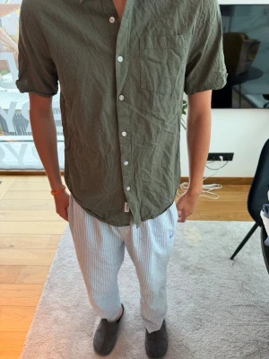 Olivgrön kortärmad skjorta - Snygg olivgrön skjorta med korta ärmar och klassisk krage. Skjortan har vita knappar framtill och en bröstficka på vänster sida. Tillverkad i ett lätt och skönt bomullstyg, perfekt för varmare dagar.