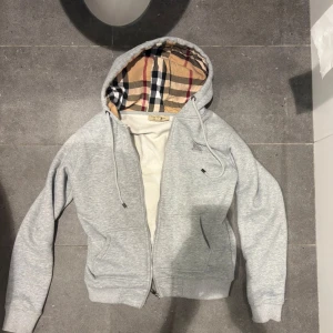 Grå hoodie från Burberry med dragkedja - Snygg grå hoodie från Burberry i storlek S med klassiskt rutigt foder i huvan. Tröjan har dragkedja framtill, snörning vid huvan och ribbade muddar. Perfekt för dig som gillar stilrena detaljer och exklusiva märken.