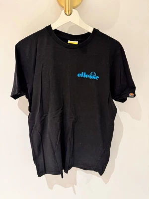 Svart t-shirt från Ellesse med tryck - Snygg svart t-shirt från Ellesse med blå logga på bröstet och stort färgglatt grafiskt tryck i rosa, blått, vitt och grått på ryggen. Klassisk passform och rund hals. Perfekt för dig som gillar streetwear och vill sticka ut lite extra.