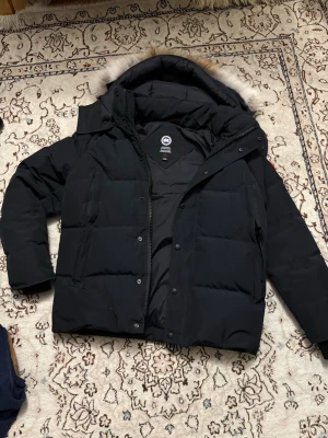 Canada goose jacks - Säljer en svart dunjacka från Canada Goose med huva och avtagbar pälskant. Jackan har quiltad design, dragkedja och knappar framtill samt lång ärm. Perfekt för kalla vinterdagar och riktigt snygg streetstil.