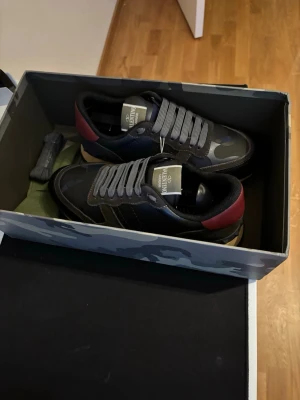 Valentino Rockrunner sneakers svart/grå - Snygga Valentino Rockrunner sneakers i svart och grått med mörkröda detaljer på hälen. Skorna har kamouflagemönster, snörning och beige sula. Tillverkade i en mix av skinn och mocka för en exklusiv känsla.