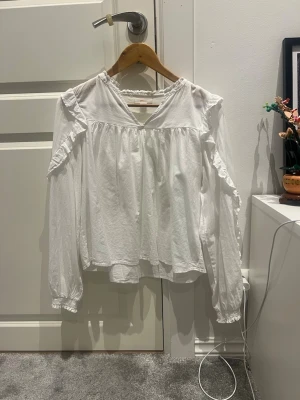 Vit volangblus med v-ringning - Söt vit blus med v-ringning och volangdetaljer längs axlarna. Blusen har långa ärmar med resår vid ärmslut och en lös, flowy passform. Perfekt för en trendig och avslappnad look.