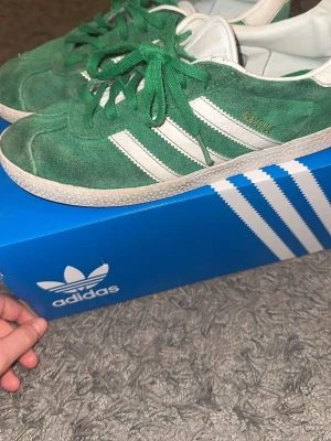 Gröna Adidas Gazelle sneakers - Gröna Adidas Gazelle sneakers i mocka med vita detaljer och klassiska tre ränder på sidan. Skorna har snörning och vit platt sula. Guldfärgad Gazelle-text på sidan och vit hälflik. Perfekta för dig som gillar retrostil och streetwear.