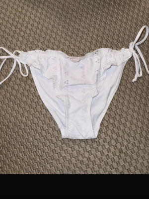 Vita bikinitrosor med broderade detaljer - Säljer ett par vita bikinitrosor med söta broderade håldetaljer och knytband i sidorna. Modellen har en feminin och romantisk look med volangkant upptill. Perfekt för strandhäng eller poolparty!