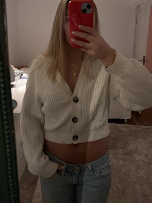 Vit cropped kofta med stora knappar - Säljer en vit, ribbstickad cropped kofta med v-ringning och stora bruna knappar framtill. Koftan har långa ärmar och en avslappnad passform, perfekt att styla med höga jeans eller kjol. Materialet känns mjukt och stretchigt.