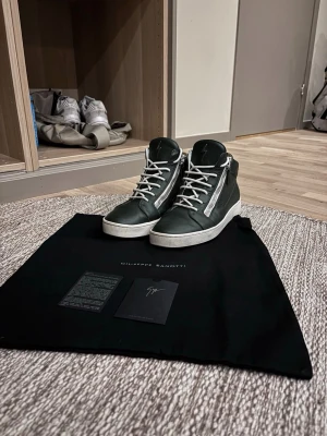 Svarta high-top sneakers Giuseppe Zanotti - Snygga svarta high-top sneakers från Giuseppe Zanotti med vita snören och dragkedjor på sidorna. Skorna har en vit platt sula och är tillverkade i skinn. Perfekta för dig som vill ha en stilren och exklusiv look. Strl 41 men passar 42 jätte bra! Har själv 42 och sitter jättebra! Nypris 5000-6000kr