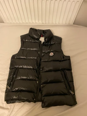 Svart dunväst från Moncler - Snygg svart dunväst från Moncler med hög krage och två dragkedjeförsedda fickor. Västen har Moncler-logga på bröstet och quiltad design med glansigt yttertyg. Perfekt för lager-på-lager och streetwear-stil.
