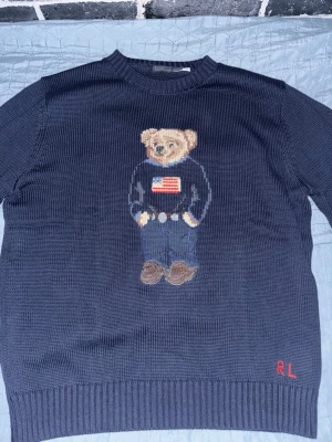 Ralph Lauren stickad tröja Polo Bear - Mörkblå stickad tröja från Ralph Lauren med den ikoniska Polo Bear på bröstet, klädd i jeans och tröja med amerikanska flaggan. RL-broderi i rött nere vid fållen. Perfekt nu i vintern och inför våren. Storlek L men passar M väldigt bra. Pris går att diskutera enbart vid snabbaffär.
