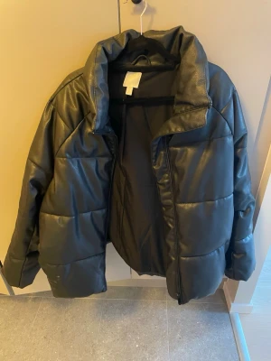 Svart quiltad pufferjacka i skinnimitation - Snygg svart pufferjacka med quiltad design och hög krage. Jackan är gjord i skinnimitation och har dragkedja framtill. Perfekt för dig som vill ha en trendig och varm jacka med lite edge.