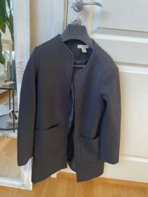 Svart kappa från H&M med fickor (obs, jag har normalt M, men denna är lite kortare i armarna) - Köpte denna på Vinted, har aldrig använt då den är lite för liten. Men det är super Stilren svart kappa från H&M i storlek 38. Modellen är rak med två stora fickor framtill och har en enkel, öppen design utan knappar eller dragkedja. Kappan har lång ärm och är fodrad för extra komfort. Perfekt för dig som gillar minimalistisk stil.