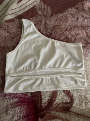 Vit oneshoulder ribbad topp - Säljer en vit ribbad topp med oneshoulder-design och en cool cut-out detalj nedtill. Perfekt för dig som gillar trendiga och unika plagg. Materialet är mjukt och stretchigt för skön passform.