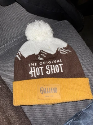 Galliano Hot Shot mössa med tofs - Snygg stickad mössa från Galliano med broderad text 'The Original Hot Shot'. Färgerna är vit, brun och senapsgul med bergsmotiv och en stor vit tofs på toppen. Märkespatch framtill. Perfekt för kalla dagar och streetstyle.