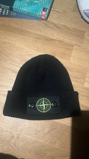 Svart ribbstickad mössa Stone Island - Svart ribbstickad mössa från Stone Island med uppvikt kant och klassisk logotyp framtill. Snygg och enkel design som passar perfekt till höst och vinter. Märket är broderat i gult och grönt på svart patch framtill. 