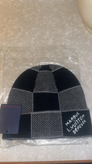 Svart rutig mössa  - Snygg mössa i svart och grått med rutigt mönster. Mössan är stickad och tillverkad av kashmir, ull och silke. Detalj med texten 'Marque L. Vuitton Depose' broderad framtill. Perfekt för dig som gillar exklusiva accessoarer.