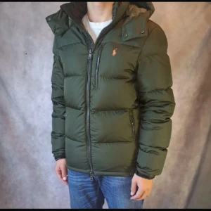 Grön pufferjacka från Polo Ralph Lauren - Snygg mörkgrön pufferjacka från Polo Ralph Lauren med huva och orange logga på bröstet. Jackan har tre dragkedjefickor fram, quiltad design och hög krage. Perfekt för kalla dagar och har en clean, modern look. Skriv vid frågor, priset går att diskuteras!