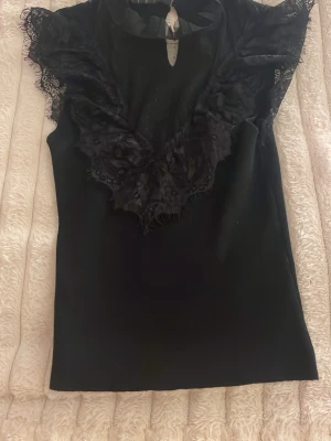 Svart spetsblus med volangdetaljer - Supersnygg svart blus med spets och volangdetaljer runt ringningen. Blusen har ärmlös design och en liten öppning framtill vid halsen. Perfekt för dig som gillar romantiska detaljer och en chic look.