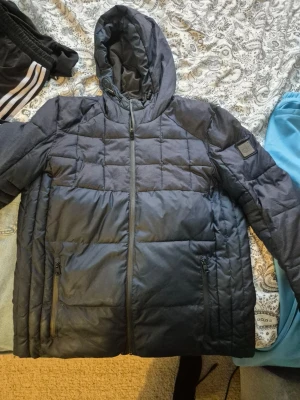 Svart pufferjacka från Calvin Klein - Säljer en svart pufferjacka från Calvin Klein med huva och dragkedja framtill. Jackan har två sidofickor med dragkedja och en snygg CK-logga på ärmen. Perfekt för kalla dagar och riktigt stilren look.