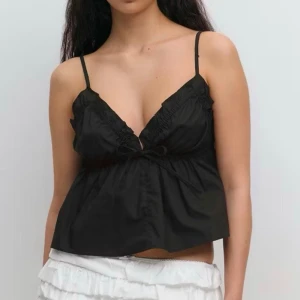 Svart linnetopp med volang och knyt - Säljer en svart linnetopp med tunna axelband, djup v-ringning och volangdetaljer längs urringningen. Toppen har en liten rosett framtill och är lätt utsvängd nedtill för en luftig känsla. Perfekt för sommarens alla tillfällen.
