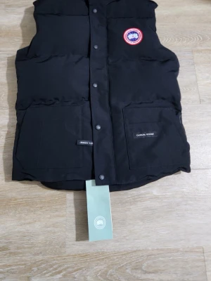 Svart dunväst från Canada Goose - Säljer en svart dunväst från Canada Goose med klassisk logga på bröstet och stora fickor framtill. Västen har tryckknappar och är perfekt för lager-på-lager under kyliga dagar. Materialet är slitstarkt och fyllningen håller dig varm.