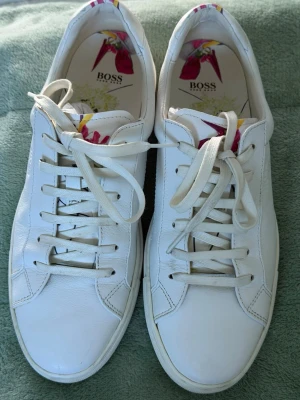 Vita sneakers från BOSS i skinn - Stilrena vita sneakers från BOSS i äkta skinn med klassisk snörning. Insidan har ett färgglatt blommönster som ger en unik touch. Perfekta för dig som vill ha en clean look med lite extra detalj på insidan.