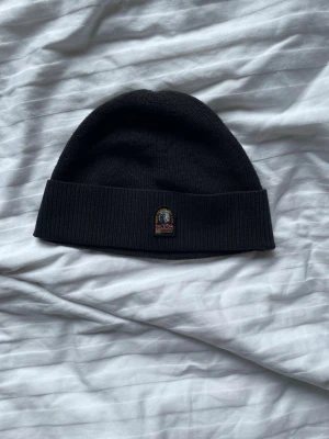 Svart mössa från Parajumpers - Svart ribbstickad mössa från Parajumpers med uppvikt kant och broderad logotyp framtill. Perfekt t vintern. Endast används fåtal gånger 
